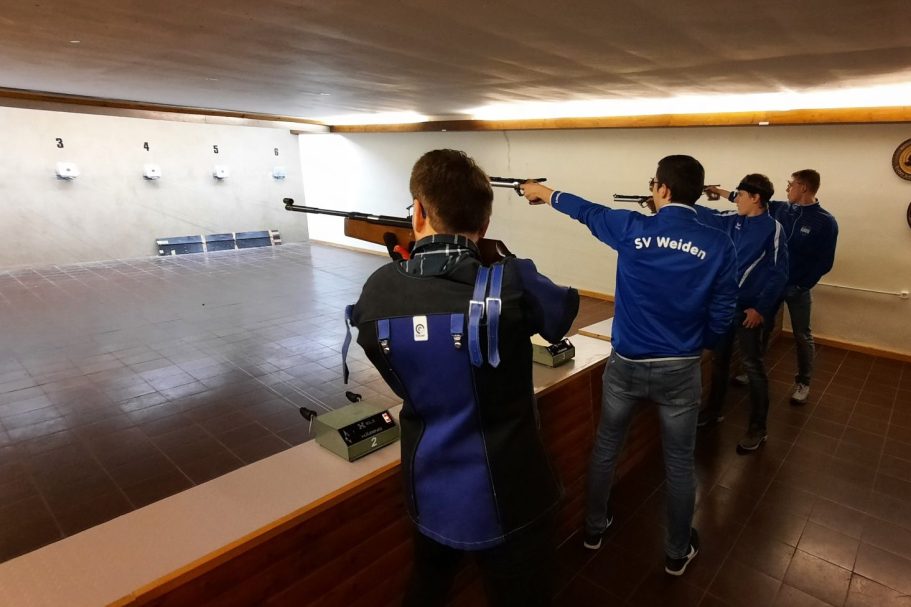 Vier Schützen in blauen Jacken, die auf Zielscheiben im Schießstand trainieren.